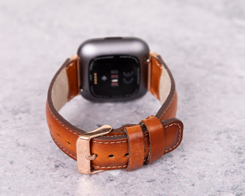 Puede incluir: Una correa de reloj de cuero marr&oacute;n con una hebilla dorada y una esfera de reloj inteligente plateada.
