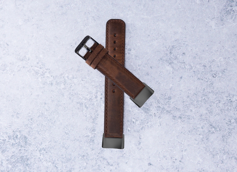 May include: A brown leather watch strap with a black metal buckle. The strap is folded in half and the buckle is visible on both ends.