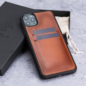 May include: A brown leather phone case with a built-in card holder. The case has a black trim and a camera cutout. The case is sitting on a gray surface.