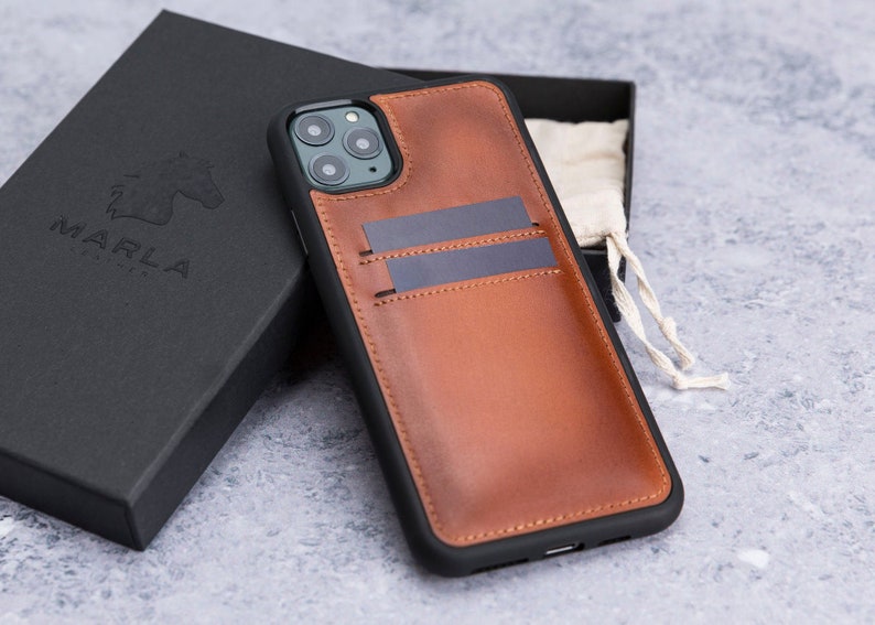 May include: A brown leather phone case with a built-in card holder. The case has a black trim and a camera cutout. The case is sitting on a gray surface.