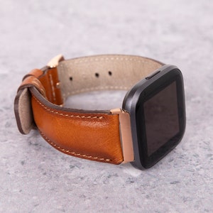 Puede incluir: Una correa de reloj de cuero marr&oacute;n con un reloj inteligente negro con una esfera cuadrada.