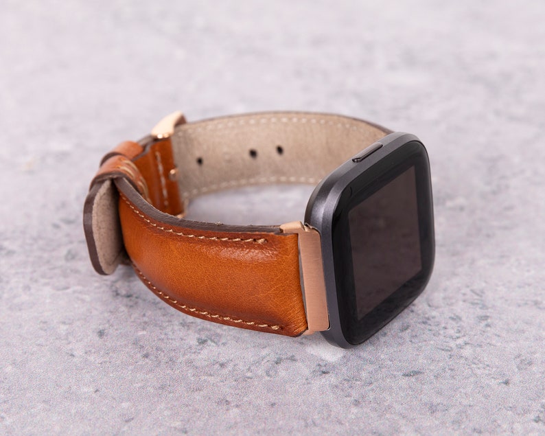 Puede incluir: Una correa de reloj de cuero marr&oacute;n con un reloj inteligente negro con una esfera cuadrada.