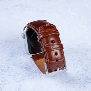 K&ouml;nnte beinhalten: Ein braunes Lederarmband mit Kroko-Muster. Das Armband ist an einer silbernen Uhr befestigt.