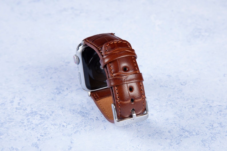 K&ouml;nnte beinhalten: Ein braunes Lederarmband mit Kroko-Muster. Das Armband ist an einer silbernen Uhr befestigt.