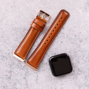 Puede incluir: Dos correas de reloj de cuero marr&oacute;n con herrajes dorados y un reloj inteligente cuadrado negro.