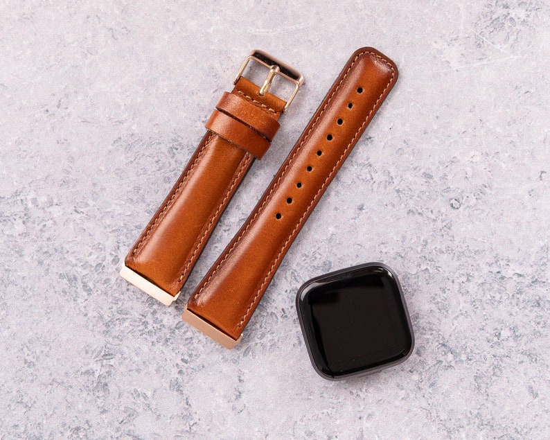 Puede incluir: Dos correas de reloj de cuero marr&oacute;n con herrajes dorados y un reloj inteligente cuadrado negro.