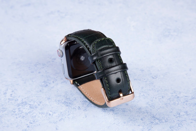 K&ouml;nnte beinhalten: Ein dunkelgr&uuml;nes Lederarmband mit einer ros&eacute;goldenen Schlie&szlig;e. Das Armband ist f&uuml;r eine Smartwatch konzipiert und hat einen gen&auml;hten Rand.