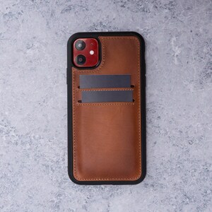 May include: A brown leather phone case with a black frame and two card slots. The case has a red camera lens cutout.