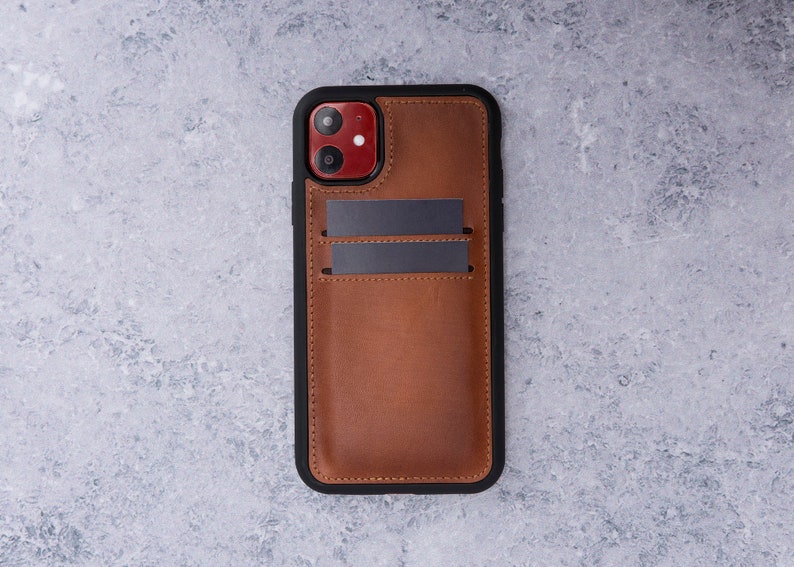 May include: A brown leather phone case with a black frame and two card slots. The case has a red camera lens cutout.