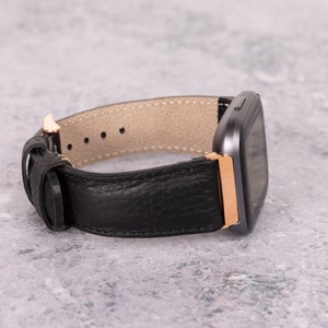 May include: A black leather watch band with a rose gold buckle and a gray square watch face.
