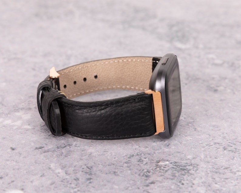 May include: A black leather watch band with a rose gold buckle and a gray square watch face.