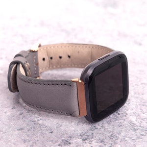 May include: A gray leather watch band with a rose gold buckle and a black smartwatch face.