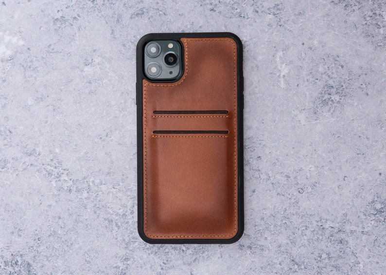 May include: A brown leather phone case with a black border and a built-in card holder. The case has a cutout for the phone's camera lens.