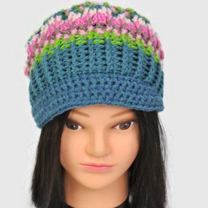 Puede incluir: Un gorro azul de crochet con rayas rosas, verdes y marrones. El gorro está en una cabeza de maniquí con cabello negro.