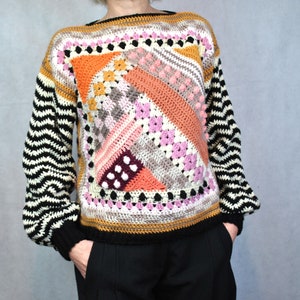 May include: A colourful crocheted sweater with a geometric pattern in shades of pink, orange, brown, and black. The sweater has long sleeves with black and white stripes.