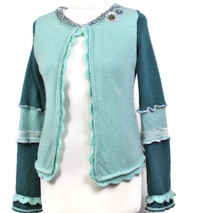 Op de afbeelding: Een teal en turquoise vest met een geschulpte rand en decoratieve versieringen. De mouwen zijn een donkerdere teal knit, en de body is een lichtere turquoise. De kraag is versierd met kralen en een bloemenpin.