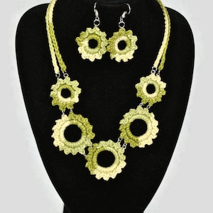 May include: A crochet jewellery set comprising a necklace and matching earrings. The necklace and earrings are made of light green and cream-coloured yarn, with a floral design. The jewellery is displayed on a black mannequin.