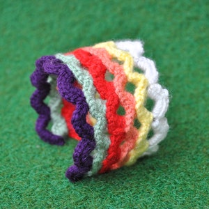 May include: A colorful, crocheted bracelet in a rainbow of hues, including purple, green, red, yellow, and white. The bracelet has a wavy, scalloped edge and is made of yarn. The craft item is displayed on a green surface.