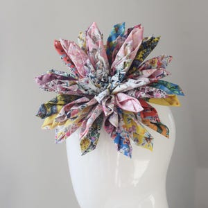 May include: A large, multi-colored fabric flower hair accessory featuring numerous folded petals in various patterns, including floral, geometric, and abstract designs in shades of pink, yellow, blue, and white, attached to a white mannequin head.