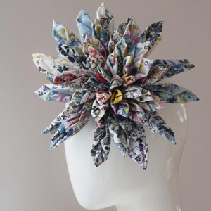 May include: A large, multi-petaled fabric flower hair accessory featuring a complex floral print in shades of blue, yellow, red, and green on a white background. The petals are folded into sharp points and accented with small, clear, sparkling rhinestones.