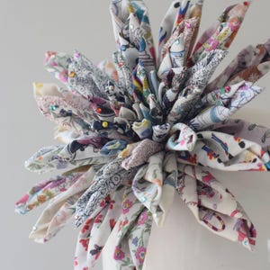May include: A decorative fabric flower sculpture constructed from numerous folded petals of white cotton fabric featuring a multi-colored floral and character print. The center is dense with layered fabric points, accented with small, colorful rhinestones.