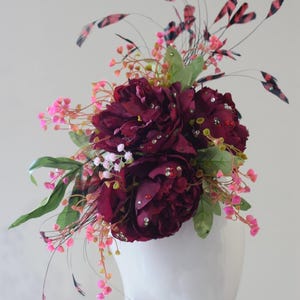 Könnte beinhalten: Burgunderfarbener floraler Kopfschmuck mit Strassakzenten, rosa und weißen Zweigen und grünen Blättern. Der Kopfschmuck hat dunkelrosa und schwarze Federn. Die Anordnung wird auf einem weißen Schaufensterkopf präsentiert.