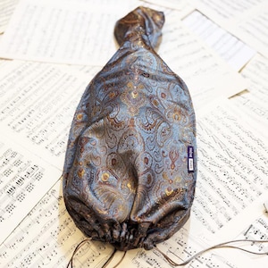 Puede incluir: Una bolsa con cordón y estampado de cachemira azul y marrón. La bolsa tiene un fondo fruncido y una parte superior retorcida. La tela tiene detalles dorados y una pequeña etiqueta que dice "IRENE PARIS". La bolsa descansa sobre un fondo de partituras.