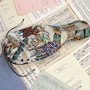 Puede incluir: Una bolsa de tela con estampado blanco y azul con cierre de cordón. La bolsa presenta una jirafa, flores y otros diseños geométricos. La bolsa está sobre partituras musicales.