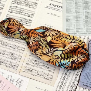 Puede incluir: Una corbata de tela marrón con un estampado repetido de hojas naranjas, amarillas y azules. La corbata se muestra sobre un fondo de partituras antiguas.