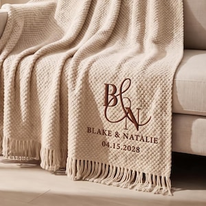 Op de afbeelding: Een crèmekleurige, getextureerde plaid met franjes, voorzien van donkerbruin borduurwerk met het monogram B&N, de namen BLAKE & NATALIE en de datum 04.15.2028. De deken is over een bank gedrapeerd en heeft een wafelachtig weefpatroon.