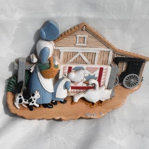 May include: A decorative figurine depicting a rural scene. It features a woman in a blue dress and bonnet, a barn, a goose, a cat, and a small buggy. The figurine has a rustic, country aesthetic with a painted finish.