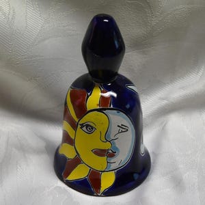 Könnte beinhalten: Eine blaue Keramikglocke mit einem bemalten Design einer Sonne und eines Mondes. Die Sonne ist gelb mit einem roten und gelben Heiligenschein, und der Mond ist weiß mit einem blauen Heiligenschein. Die Glocke ist ein dekoratives Element.