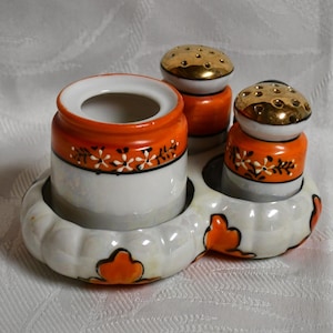 Pode incluir: Um conjunto vintage de saleiro e pimenteiro em cerâmica com um recipiente a condizer. O conjunto apresenta uma base branca com detalhes em laranja e tampas douradas. As peças são decoradas com padrões florais.