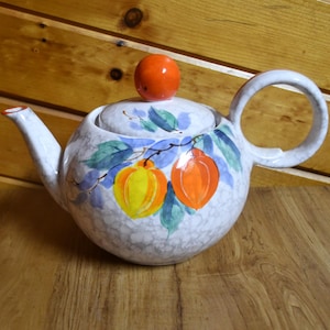 Puede incluir: Una tetera de cerámica blanca con un diseño pintado a mano de fruta naranja y amarilla en una vid. La tetera tiene un cuerpo redondo y un asa curva. La tapa tiene un pequeño pomo naranja.