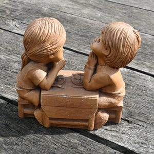 May include: A brown ceramic figurine of two children seated at a small table. The children are facing each other, with their hands near their faces. The table has two small cups on top. The figurine is on a weathered wooden surface.