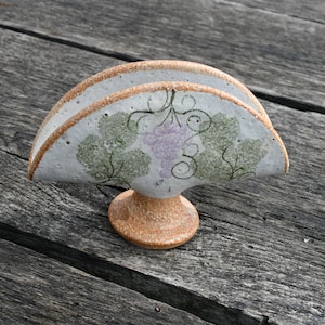 May include: A ceramic napkin holder with a pedestal base. The holder is off-white with a brown rim and features a painted design of green leaves and purple grapes. The holder is on a weathered wooden surface.