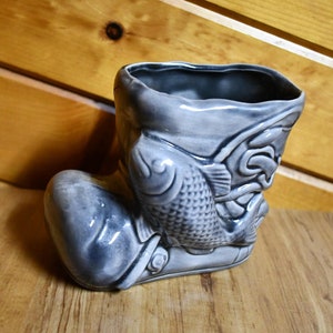 Puede incluir: Un macetero de cerámica gris en forma de bota con un diseño de pez. La bota está decorada con un pez y otros detalles.