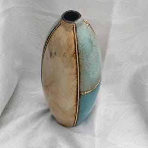 May include: An oval-shaped ceramic vase featuring a segmented design with a smooth beige section and a contrasting light blue and teal section. The segments are separated by thin, dark brown vertical and horizontal lines with a glossy finish.