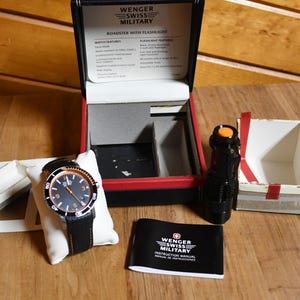 May include: A Wenger Swiss Military Roadster watch with a black dial and strap, presented on a white cushion. The set includes a black flashlight, a black and red storage box with product text, and an instruction manual. The box lid interior lists watch and flashlight features.