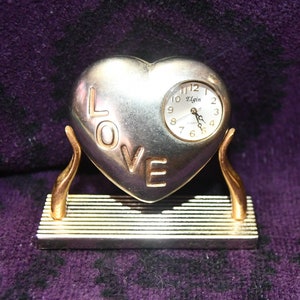 May include: A silver heart-shaped clock with the word "LOVE" embossed on the front. The clock face is white with black numbers and hands, and it says "Elgin" at the top. The clock is on a gold stand.