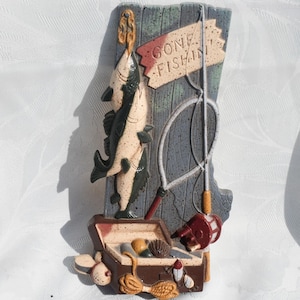 May include: A decorative wall hanging featuring a fishing theme. Two fish are suspended from a wooden sign that reads "Gone Fishin'". A fishing rod, reel, and tackle box are also included in the design. The color palette includes shades of brown, blue, and red.