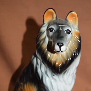 May include: A ceramic figurine of a Collie dog. The dog is sitting with its head tilted slightly to the side. It has black, white, and brown fur.