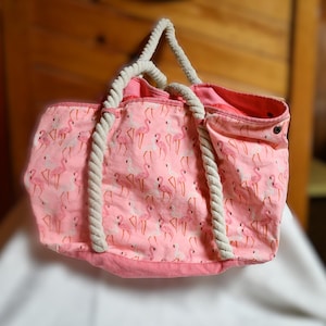 May include: Pink canvas tote bag with white rope handles and a flamingo print.