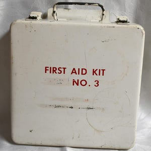 Peut inclure: Une trousse de premiers secours vintage en métal blanc avec une poignée et des loquets noirs. Les mots "FIRST AID KIT NO. 3" sont imprimés en rouge sur le devant. La trousse montre des signes d'usure.