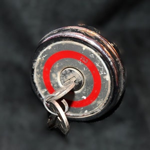 May include: A silver keyhole with a red ring around it. The keyhole is on a black metal cap with a silver rim.