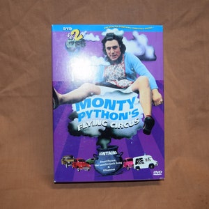 May include: DVD box set of Monty Python's Flying Circus, featuring a purple background with a man in a dress sitting on a cloud. The text "Monty Python's Flying Circus" is in white letters. The box set contains two DVDs and includes episodes like "Dead Parrot" and "The Lumberjack Song".