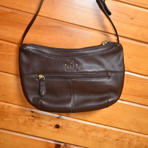 May include: A brown leather shoulder bag with a long strap, hanging against a wooden background. The bag has a zippered main compartment and a smaller front pocket, both with gold-colored zippers. The brand name "Stone Mountain" is visible on the front.