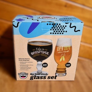 May include: A box for a set of two beer glasses. The glasses are clear and have funny sayings on them. The glass on the left says "Life is Brewtiful" and the glass on the right says "IPA Lot When I Drink". The box is blue and white with a cartoon mouth on it.