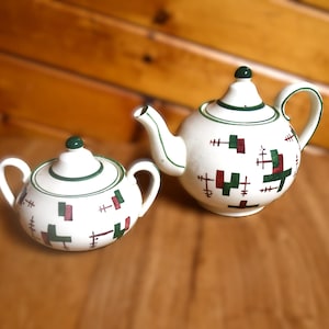 Pode incluir: Um conjunto de bule e açucareiro de cerâmica branca. O bule tem um bico e uma alça, e o açucareiro tem duas alças. Ambas as peças são decoradas com formas geométricas verdes e castanhas e acabamentos verdes.