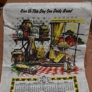 May include: A vintage kitchen towel with a calendar for the year 1974. The towel features a colorful illustration of a kitchen scene with jars, a pitcher, a loaf of bread, and a window with a view of a red barn. The text "Give Us This Day Our Daily Bread" is printed at the top of the towel.